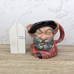 Op de afbeelding: Een keramische kan met een lachend gezicht, een zwarte hoed en een lange grijze baard. De kan is rood, zwart en bruin. De kan is ongeveer 15 cm hoog. Naast de kan staat een wit houten huisje.