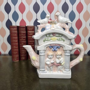 May include: A decorative ceramic teapot with a whimsical design. The teapot features a white structure with two cherubic figures, a roof with two doves, and a pink and yellow handle and spout. The teapot is approximately 20 cm tall.