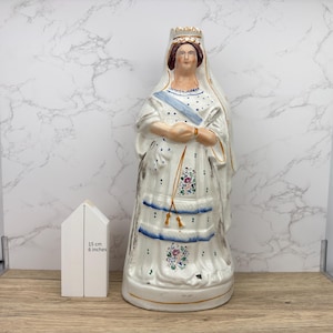 May include: A white ceramic figurine of a woman wearing a crown and a long white dress with blue and gold accents. The figurine is 6 inches tall.