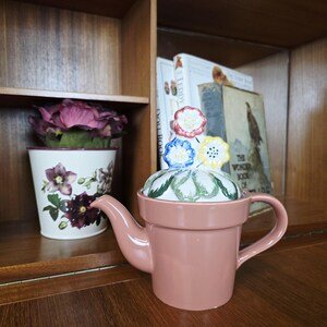May include: A pink ceramic teapot with a flower-shaped lid. The lid features red, blue, and yellow flowers. The teapot is in the shape of a flower pot. A decorative flower pot with purple flowers is in the background.