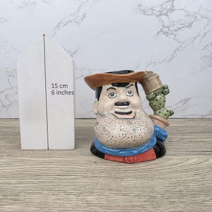 以下が含まれることがあります： 口ひげとあごひげを生やし、帽子をかぶりサボテンを持っている男性の形をしたセラミックノベルティピッチャー。ピッチャーの高さは約15cmです。男性の服装には、青いスカーフと赤いアクセントが含まれています。