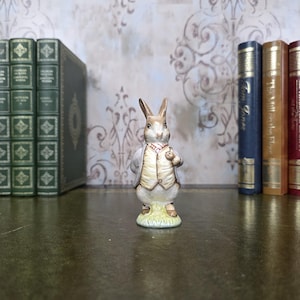 May include: A ceramic rabbit figurine, dressed in a waistcoat and jacket, holding a pocket watch. The rabbit stands on a patch of grass. The figurine is approximately 10 cm tall.
