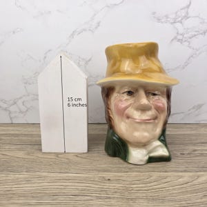 Puede incluir: Una taza de cerámica con forma de rostro de hombre sonriente, con sombrero amarillo y chaqueta verde. La taza está junto a un objeto blanco con forma de casa. El objeto con forma de casa mide 15 cm de alto.