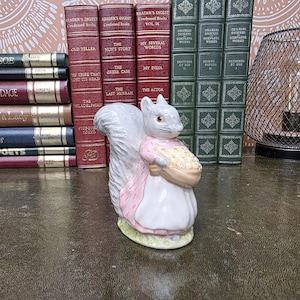 Könnte beinhalten: Eine stehende Eichhörnchen-Figur aus Keramik, die ein hellrosa Kleid trägt und einen Korb mit kleinen, runden, hellen Gegenständen hält. Das Eichhörnchen hat einen grauen Schwanz und steht auf einem kleinen, ovalen Sockel mit hellgrüner Textur.