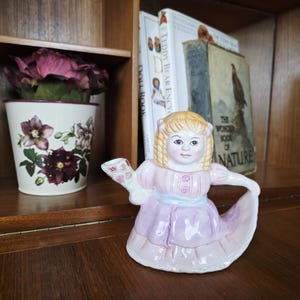 May include: A ceramic girl figurine in a pink and lilac dress, holding flowers. The figurine has blonde hair and is on a wooden surface. A floral pot and books are in the background, creating a vintage aesthetic.