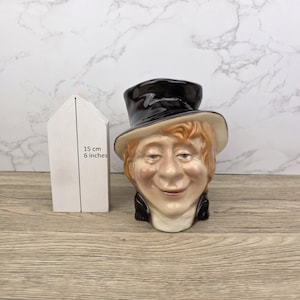 May include: A ceramic character jug with a smiling face, wearing a black top hat and a cream-colored brim. The jug is approximately 6 inches tall. A white box is next to the jug, with the dimensions of the jug labeled on the side.