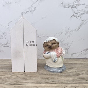 以下が含まれることがあります： 帽子とエプロンを着て白い布を持っているハリネズミの陶器製フィギュア。フィギュアは灰色の台座にあります。隣にある白い木製の家の形は高さ15cm（6インチ）です。