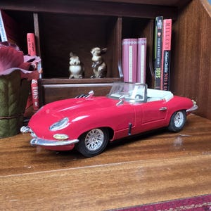 以下が含まれることがあります： 白い内装の赤いおもちゃのコンバーチブルカー。車は銀色のホイールで、木製の表面にあります。車の後ろには、セラミックのウサギの置物と、「TOYS」と「MILLER'S Antiques Checklist TOYS & GA」というテキストが書かれた本があります。