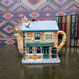 Puede incluir: Una tetera de cerámica con forma de la posada Rovers Return Inn, con fachada de ladrillo color canela, adornos verde azulado y techo gris. El letrero dice Rovers Return Inn y Newton and Ridley Best Bitter. Un gato blanco y negro está sobre el tejado.