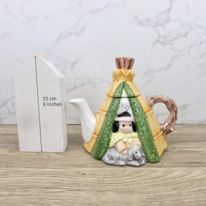 Pode incluir: Uma chaleira de cerâmica em forma de tipi nativo americano, com uma figura e uma ovelha. A chaleira é amarela, verde e castanha, com um bico e uma pega brancos. A altura é de 15 cm.