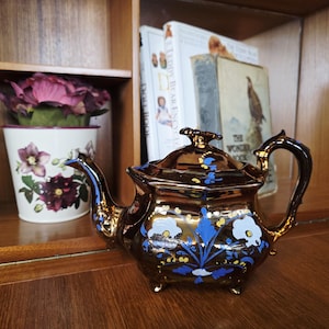 Puede incluir: Una tetera marrón decorativa con detalles dorados y diseños florales azules. La tetera tiene un pico y un asa curvados, y está sobre una superficie de madera. Un jarrón floral y libros están en el fondo.