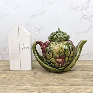 Può includere: Teiera in ceramica con un design a carciofo verde, rosso e giallo. La teiera è alta circa 15 cm. Il coperchio è anch'esso verde e a forma di foglia. Il manico e il beccuccio sono curvi.