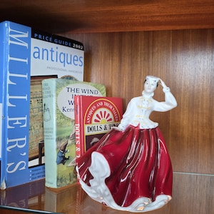 May include: A porcelain figurine of a woman in a long red skirt and white jacket with gold trim. The figurine is standing on a shelf next to several books, including a price guide for antiques and a book titled "Dolls & Toys."