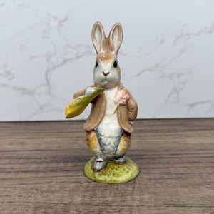 May include: A ceramic rabbit figurine wearing a brown jacket and holding a leaf. The rabbit has a pink flower on its lapel and stands on a green base. The figurine is painted with detailed features, reminiscent of a children's storybook character.