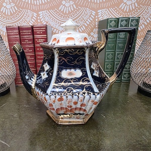 Op de afbeelding: Een zeshoekige keramische theepot met een diep marineblauw en wit kleurenpalet met ingewikkelde gouden filigraanaccenten. De behuizing is versierd met oranje bloemmotieven en geschulpte patronen, aangevuld met een bijpassend deksel met knop.