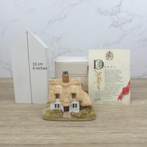 May include: A miniature cottage figurine with a thatched roof, two windows, and a chimney. The figurine is approximately 6 inches tall. A white box and a card with text are in the background.