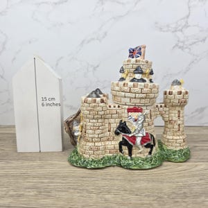 May include: A ceramic castle ornament featuring a knight on horseback, soldiers, and a Union Jack flag. The beige castle has brick detailing and green grass at the base. The ornament is approximately 15 cm tall.