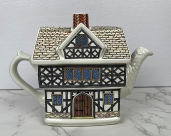 Puede incluir: Una tetera de cerámica con forma de casa estilo Tudor de dos pisos con entramado de madera negro, paredes blancas, ventanas azules, una puerta de madera marrón y un techo texturizado. El asa está a la izquierda y un pico blanco a la derecha.