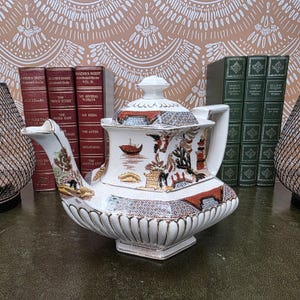 Op de afbeelding: Een witte keramische theepot met een vierkante voet en deksel, voorzien van gedetailleerde, veelkleurige oosterse illustraties van pagodes, bruggen en boten. De theepot heeft een geribbelde onderkant, een tuit en een handvat met gouden rand.