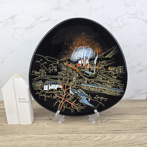 May include: Black decorative plate with a landscape scene in gold and orange tones. The plate features a townscape with buildings, trees, and the word "Longwy". The plate is 6 inches in diameter.