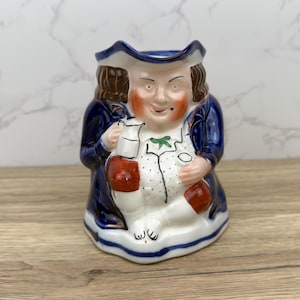 May include: A ceramic pitcher in the shape of a man wearing a blue coat, white shirt, and a brown wig. He is holding a small pitcher in his right hand and has a green bow tie.