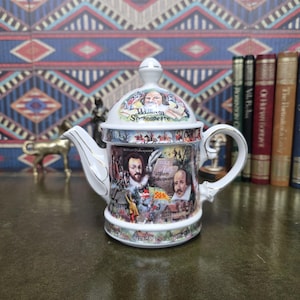 Könnte beinhalten: Eine weiße Keramik-Teekanne mit farbenfrohen Illustrationen von William Shakespeare, historischen Szenen und dem Schriftzug William Shakespeare. Deckel und Körper sind mit detaillierten Porträts und Landschaften verziert, akzentuiert durch Goldränder an Griff und Ausguss.