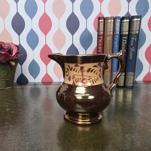 May include: A ceramic pitcher with a metallic bronze finish, featuring a cream-colored band around the upper body decorated with dark brown leaf-like patterns. The pitcher has a curved handle, a flared spout, and a circular base.