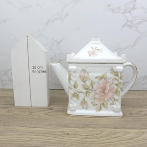 May include: White ceramic teapot with a square shape and a floral design in pink and green. The teapot has a gold handle and trim. A white wooden house shape is next to the teapot, with the text "15 cm 6 inches".