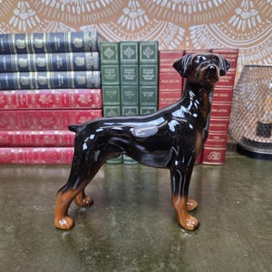 May include: A glossy ceramic figurine of a Doberman Pinscher dog with a black and tan coat. The dog is standing in a profile position, facing right. A small oval sticker on the side of the dog's body reads COOPERCRAFT MADE IN ENGLAND.