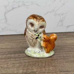 May include: A ceramic figurine of a brown and white owl with a brown squirrel beside it. The owl is holding a green twig in its beak. The figurine is on a green base.