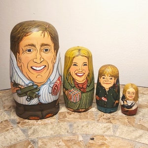May include: Set of four hand-painted wooden nesting dolls, or matryoshka dolls, featuring portraits of a family. The largest doll is a man with brown hair and a blue and white striped shirt. The second largest doll is a woman with blonde hair and a green dress. The third largest doll is a boy with blonde hair and a green jacket. The smallest doll is a girl with blonde hair and a pink dress.