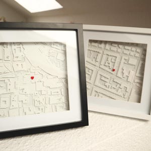 Könnte beinhalten: Zwei gerahmte Stadtkarten-Kunstwerke. Ein Rahmen ist schwarz, der andere weiß. Jede Karte ist ein detailliertes, geschichtetes Papier-Schnitt-Design mit einem roten Herzen, das einen Ort markiert.