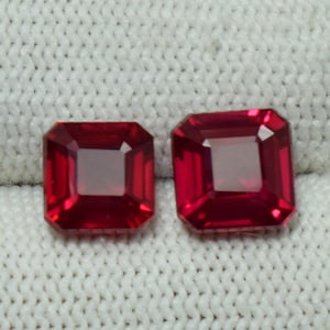 Op de afbeelding: Twee vierkant geslepen, levendige rode edelstenen. Elke edelsteen heeft een gefacetteerd oppervlak dat licht reflecteert. De edelstenen worden gepresenteerd op een wit, getextureerd oppervlak, dat hun kleur en helderheid benadrukt. Ideaal voor het maken van sieraden.