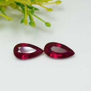 Könnte beinhalten: Zwei birnenförmige, facettierte, tiefrote Edelsteine. Die Edelsteine werden auf einer weißen Oberfläche präsentiert, mit einem Zweig gelb-grünen Laubes im Hintergrund. Die Edelsteine haben einen hohen Glanz und sind wahrscheinlich Rubine.