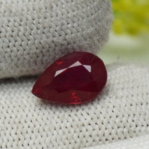 Peut inclure: Une pierre précieuse en forme de poire, taillée en facettes, de couleur rouge foncé. La gemme présente des facettes brillantes et réfléchissantes. Le rubis est posé sur un fond gris clair texturé.