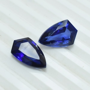 Puede incluir: Dos gemas facetadas de color azul intenso con una forma única de escudo. Las gemas están talladas con múltiples facetas, reflejando la luz y creando un brillo brillante. Las piedras están sobre un fondo blanco.