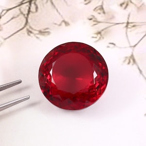 May include: A round, faceted, deep red gemstone with a brilliant cut. The stone is positioned next to a pair of silver-toned metal tweezers, highlighting the reflective surface and the precise geometric facets of the circular red gem.