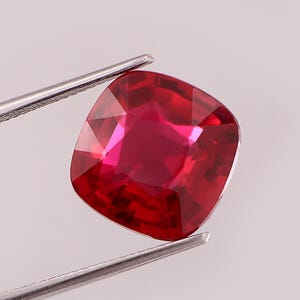 Pode incluir: Uma gema vermelha de corte almofada com uma superfície facetada que reflete a luz, mantida por uma pinça de metal prateado. A pedra exibe um tom carmesim profundo e saturado com clareza interna clara e bordas bem definidas.