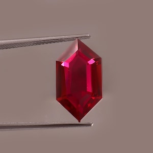 Peut inclure: Une pierre précieuse rouge vif, taillée en forme hexagonale. Tenue par une pince argentée, ses facettes captent la lumière, révélant sa clarté et sa couleur intense. Le fond est neutre. Cette gemme est probablement destinée à la joaillerie.