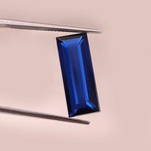 Puede incluir: Una gema rectangular de color azul oscuro es sostenida por unas pinzas plateadas sobre un fondo rosa suave. La gema tiene un corte facetado, que refleja la luz y muestra su rico color. Las pinzas están posicionadas para mostrar la gema.