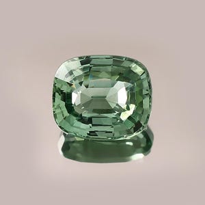 Puede incluir: Una gema verde claro, talla cojín, con una superficie reflectante. La gema se presenta sobre un fondo neutro, resaltando sus facetas y claridad. El color de la gema es un verde suave y acogedor.