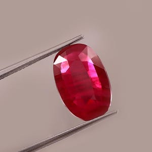 May include: A vibrant, oval-cut ruby gemstone held by tweezers. The ruby displays a deep red hue with internal light reflections. The gemstone is a focal point, showcasing its polished facets and clarity.