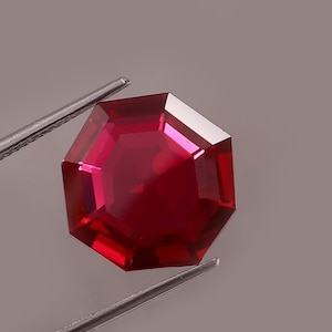 Puede incluir: Una gema facetada de color rojo intenso con una forma geométrica de múltiples lados. La gema está sujeta por unas pinzas plateadas sobre un fondo gris neutro. Las facetas de la gema reflejan la luz, creando una apariencia brillante.