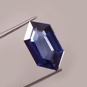 May include: A deep blue gemstone, rectangular with angled edges, held by silver tweezers. The polished surfaces reflect light, highlighting the gem's internal structure. The colour varies from dark indigo to lighter shades, showcasing its clarity.