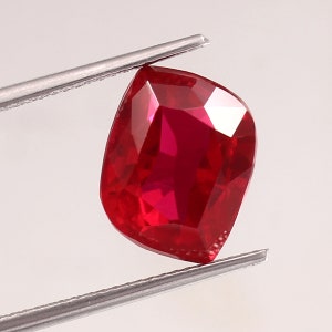 Op de afbeelding: Een gefacetteerde robijn edelsteen in kussenvorm, vastgehouden door een metalen pincet. De robijn heeft een diepe, verzadigde rode kleur, met meerdere facetten die het licht reflecteren. De edelsteen staat op een witte achtergrond.
