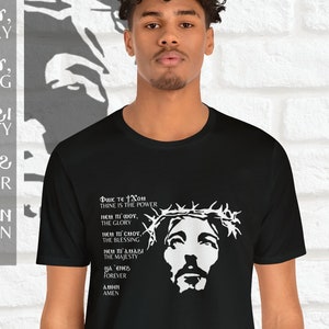 May include: Black t-shirt with a white image of Jesus Christ wearing a crown of thorns. The text on the shirt reads "Φωκ τε Χου Thine is the Power ΝΕΜ ΠΙ ΜΟΥ, The Glory ΝΕΜ ΠΙΣΜΟΥ, The Blessing THE MAJESTY WA ENE FOREVER ΑΜΗΝ AMEN".