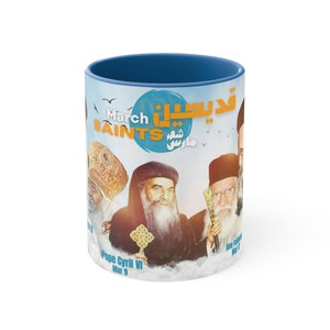 May include: A white ceramic mug with a blue rim. The mug features a colourful design with images of religious figures and the text "March Saints" and "قديسينا".