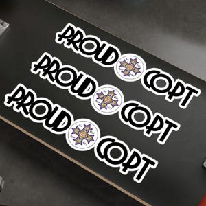 May include: White stickers with the text "PROUD COPT" in a bold, black font. Each sticker features a circular emblem with a cross design in yellow, blue, and purple. The stickers are arranged on a black surface.