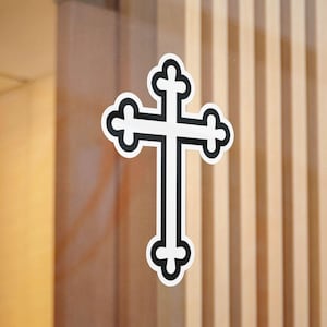 May include: A white cross with a black outline and decorative ends. The cross is affixed to a glass surface, with a wooden background visible. The cross is a symbol of faith and spirituality.