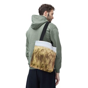 Könnte beinhalten: Eine Person in einem grünen Kapuzenpullover trägt eine schwarze Tragetasche mit einem gedruckten Bild eines goldenen Weizenfeldes.